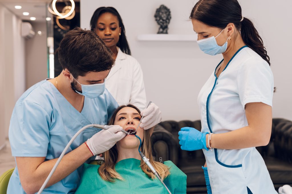 Attractive young woman in stomatology clinic being examined by a male dentist. Healthy teeth concept. Oral hygiene and dental care. Doctor fixing patient teeth with a help of a two assistant.