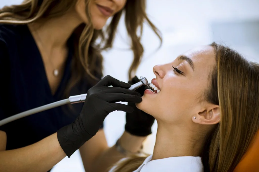 Dentist in black scrubs and gloves performs a routine dental cleaning on a relaxed patient in a bright, modern clinic with professional soft lighting.