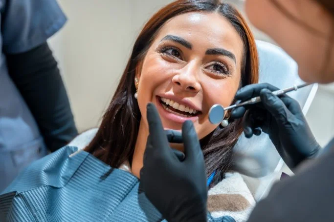Smiling woman in a dental chair receiving a checkup from a practitioner wearing black gloves under bright clinical office lighting.