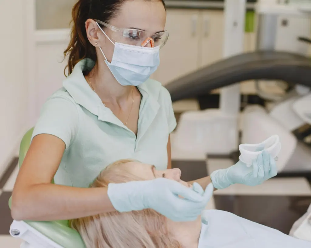Dentist in scrubs, mask, and safety glasses examines a patient in a bright, modern clinic, holding a dental tray in gloved hands.