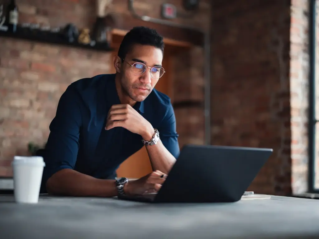 Focused professional in glasses working on a laptop in a warm-lit, brick-walled office with a coffee cup on the desk.