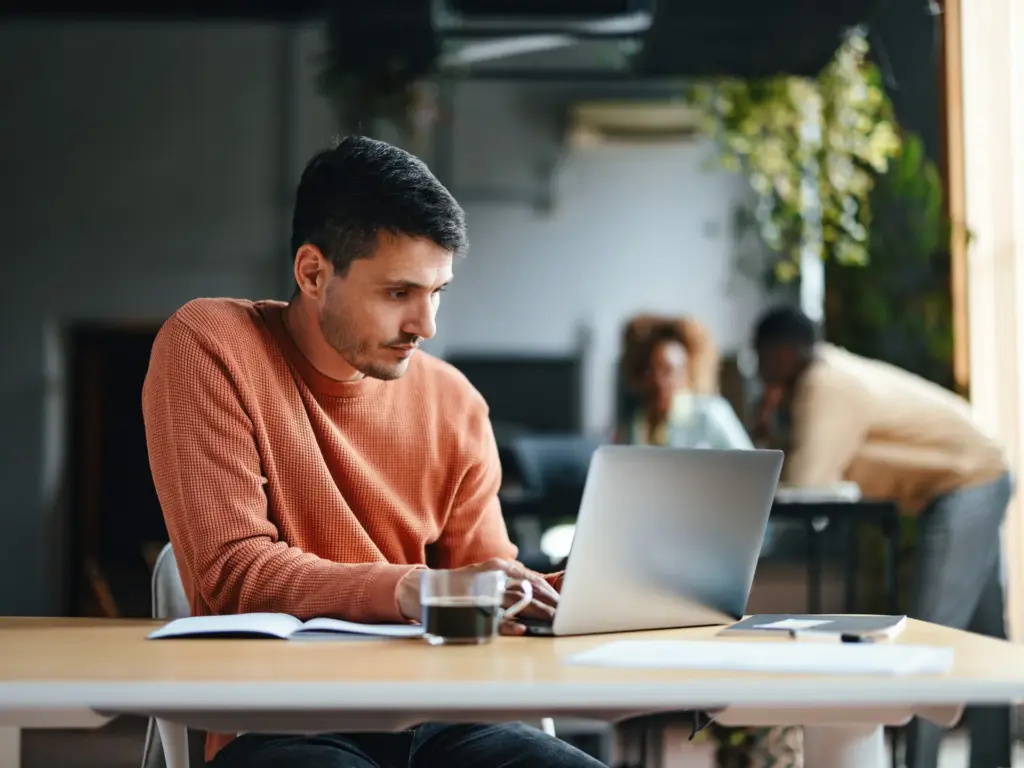 Man in rust-colored sweater working on silver laptop in modern, naturally lit office with open-plan workspace and blurred coworkers in background.