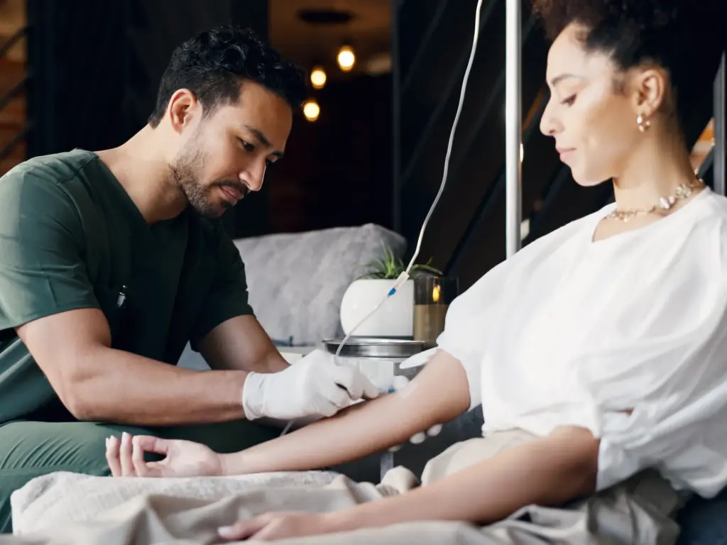 Male nurse in green scrubs administers IV therapy to a female patient in a modern, warm-lit wellness clinic setting with soft ambient lighting.