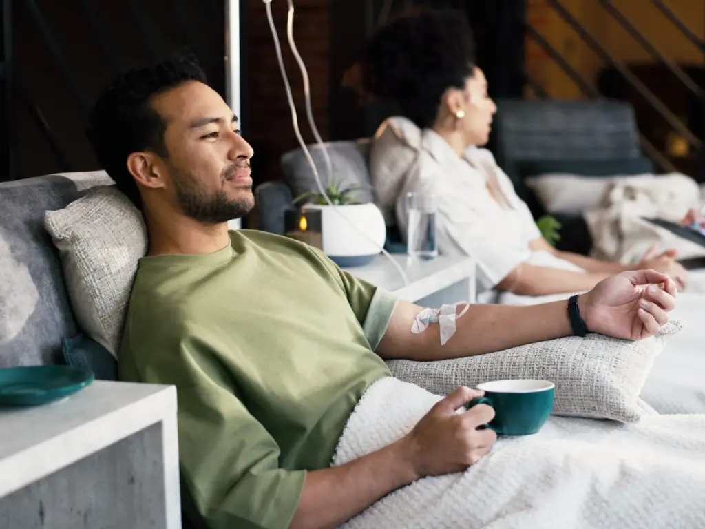 Man in olive shirt relaxing with IV drip therapy in a softly lit, modern lounge. Beside him, a woman rests in a plush chair with a drink.
