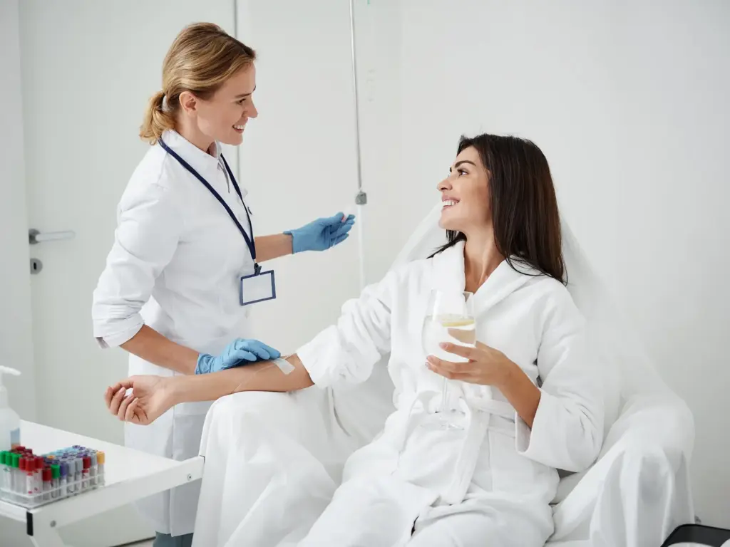 Smiling nurse in a white coat administering an IV drip to a woman in a white robe in a bright, modern, clinical medical spa setting.