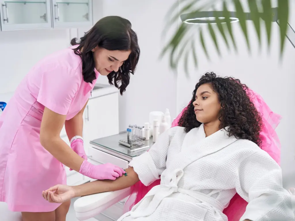 Practitioner in pink scrubs administers an IV drip to a patient in a white robe in a brightly lit, professional medical aesthetics clinic.