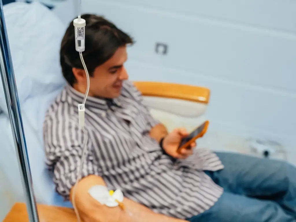 Patient receives IV therapy in a brightly lit clinic, smiling while checking their smartphone. Close-up on the IV drip set and infusion site.