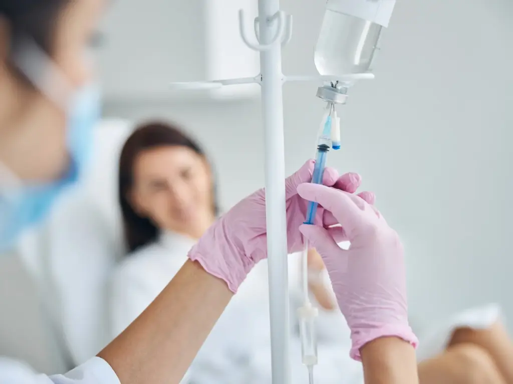 Nurse in pink gloves adjusts an IV drip for a patient in a brightly lit medical room, focusing on the sterile setup and saline bag equipment.